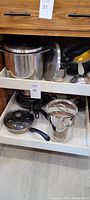 Large stainless steel pots and frying pans stored in kitchen cabinet drawer, including a strainer with a yellow handle.