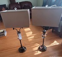 Two living room lamps side by side showing beige rectangular shades and bronze/chrome bases with crystal finials on a hardwood floor.
