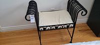 Front view of black metal bedroom bench with cream cushion on wooden floor.