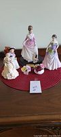 Full lot layout showing three Royal Doulton female figurines on a red placemat with three small porcelain flower arrangements in front.