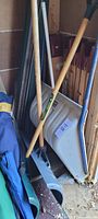 Photo showing three shovels resting upright, including a white plastic scoop shovel and two wooden handled shovels.