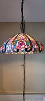 Full view of stained glass pendant lamp hanging from ceiling, colorful floral pattern visible, lamp turned on producing warm light.