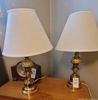 Two brass lamps side by side on a wooden surface, showing different beige lamp shades and polished brass bases.