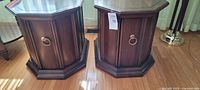 Two dark wood octagonal end tables side by side on a wood floor, showing the front with cabinet door and brass circular handle