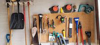 Wide angled photo showing pegboard wall with various gardening and garage hand tools including shovels, rakes, saws, pruning shears, and measuring tapes.
