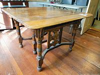 Oblique front angle of the antique wooden square dining table showing decorative apron and turned legs on hardwood floor.