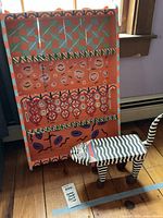 Front view of the hand painted wall shelf and small cat stool on the floor near a window, showing the colorful patterns and striped stool