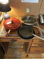 Four frying pans on a wooden desk: red enameled, two black non-stick, and one stainless steel with glass lid