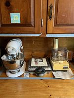 Wide shot showing all three appliances on counter under wood cabinets: white KitchenAid mixer on left, Villaware waffle maker center with manual on top, yellow Cuisinart food processor on right with manual below.