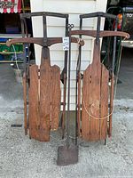 Front view and overview of two antique Flexible Flyer sleds side by side with rustic metal frames and wooden slats, accompanied by a primitive hand forged wrought iron fireplace shovel