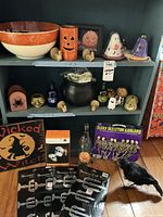 Photo shows shelves with various Halloween decorations including orange and black ceramic bowl, ceramic jack-o-lantern vase, ghost figurine, turkey figurines, green skull, crow figurine, skeleton garland package, Halloween wall decals, and Halloween-themed signs and salt and pepper shakers.