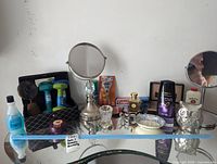 Collection of vanity and beauty items arranged on glass shelf, including mirrors, perfumes, shampoo, weights, razor, and cream.
