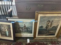 Photo showing three framed colonial prints including the hunter, equestrian Charles Davis, and domestic scene, positioned on floor against an antique chest of drawers.