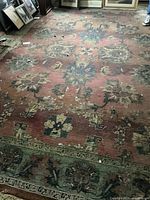 Photo showing the full rug with its worn, weathered condition and floral tapestry pattern in dark tones, including some holes and dirt.