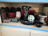 Shelf view showing carved wood plaques, red lacquer bowls, cloisonné-style box, square planter, white pierced vase, spongeware pieces, green orb, red panel and blue pedestal with bird