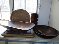 Photo showing large wooden bowls, wooden spoon, granite cutting boards, large wooden cutting board, and woven straw placemat with natural light background by a window.