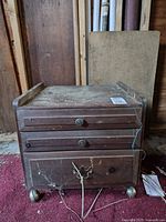 Front view of the heavily used vintage wooden sewing cabinet on casters with three drawers and metal bottom latch, showing dust and cobwebs
