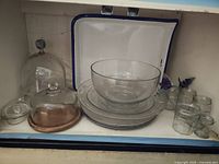 Photo showing large glass bowl, stack of plates, glass cloches (including one with wooden base) and six small jars all arranged inside a white cabinet.