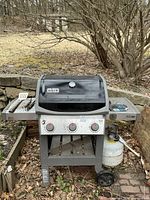 Full frontal view of the Weber grill showing three control knobs, propane tank attached, grill spatula and tongs on left shelf.