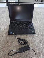 Lenovo ThinkPad laptop open with screen, keyboard visible, charger cable shown on carpet.