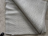 Close-up photo of folded rug showing its woven textured geometric diamond pattern in neutral tones.