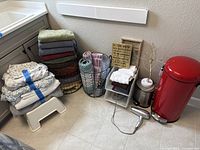 Multiple household items on tiled floor including towels, laundry baskets, decorative frames, red trash bin, small silver trash bin, hair dryer, gold decorative tree with USB cable.