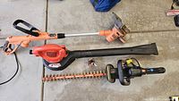Full view of the three tools placed on garage floor: Black and Decker weed wacker with pole extender, Black and Decker leaf blower, and Remington hedge trimmer with visible orange and black housing.