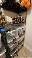 Black metal shelving unit seen with multiple adjustable wire mesh shelves holding plastic bins and toy truck. Shelving is positioned in room corner on carpeted floor.