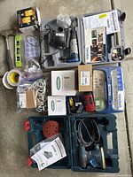 Overview of all items arranged on the garage floor showing Bosch palm sander case, Dremel case, light bulbs, tape, ax, small level, curtain rod holders, and instruction manuals.