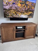Front view of the walnut entertainment system under a mounted TV, showing the cabinet with closed wood doors on each side and open middle section with shelving and electronic devices.