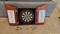 Full view of closed dartboard cabinet open showing dartboard, scoreboards on doors, six darts and marker on floor.
