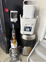Full view showing Dyson vacuum, two white Igloo coolers stacked, and silver trash can with black liner bag beside stairs.