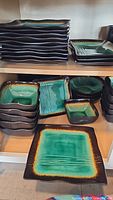 Stack of large square dinner plates and salad plates on kitchen shelf, green and black ceramic dishware.