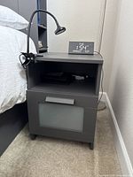 Full view of the dark gray nightstand next to bed, showing digital alarm clock on top and desk light clamped to side.