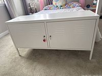 Front view of white metal storage cabinet with two lockable doors on beige carpet near bed.