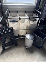 Overview photo showing laundry sorter with white cloth bags, folding step stool, trash bin, and recycling bin next to window.