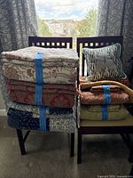 Two stacks of folded bedding on chairs by a window: three larger sets on left, smaller items and throw pillow on right.