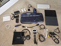 Main overview of all items arranged on carpet: includes keyboards, tablet, mouse, headphones, tripod with ring light, camera, chargers and cables