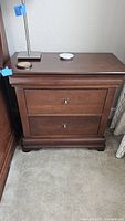 Front view of a solid wood end table/nightstand with dark brown finish. Shows two brass hardware handles and closed drawer fronts.