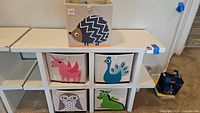 White wood shelving unit showing cubbies with five animal-themed canvas storage bins including hedgehog, unicorn, peacock, owl, and dragon designs