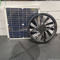 Full view showing the 100W solar panel, 10-inch window fan, and metal fan guard together on a black surface.