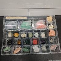 Top view of kit tray showing various gemstones and crystals neatly placed in individual compartments, along with clear plastic cups filled with colored powders/substances and some small bags.