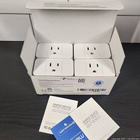 Open box showing 4 white Meross MSS110 smart WiFi outlet plugs inside with instruction manuals on a black surface.