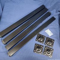 Four matte black square steel table legs laid on a blue fabric background with four square mounting plates and a bag of screws.