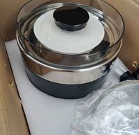 Stainless steel inner pot with transparent lid and black knob, white strainer, and power cord inside packaging box.