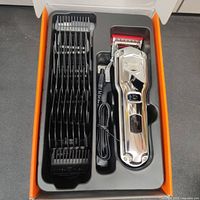 Open box showing hair clipper, multiple guide comb attachments, and charging cable in tray.