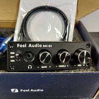 Front view of Fosi Audio DAC-Q4 USB DAC headphone amplifier showing USB, optical, coaxial inputs switch, headphone jack, bass, treble and volume knobs