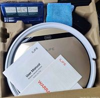 Shot shows ILIFE V5s Plus robot vacuum and mop in packaging with user manual, warranty papers, charging adapter, cleaning cloth.