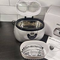 Front and top view of ultrasonic cleaner open with white basket placed beside it, showing digital timer display and open lid with transparent window.