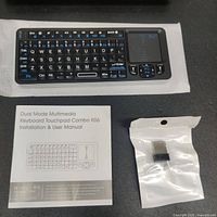 Mini wireless Bluetooth keyboard with backlit keys and touchpad laid flat on a surface next to USB receiver and user manual.